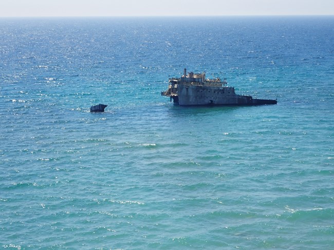 Two gray-colored parts of a shipwreck are visible above the surface of blue water.