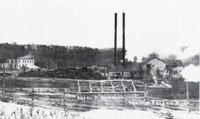 large sawmill with two tall smokestacks protruding from the rood sits next to a large pile of cut logs. Behind it sits a square, two-story hotel and other wooden buildings. Text across the bottom reads: “Hotel + sawmill, Manitou Island, Mich.”