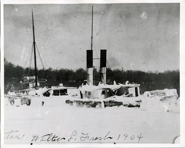 Black and white photo of a steam-powered vessel trapped in thick ice, surrounded by snow-covered terrain and bare trees.