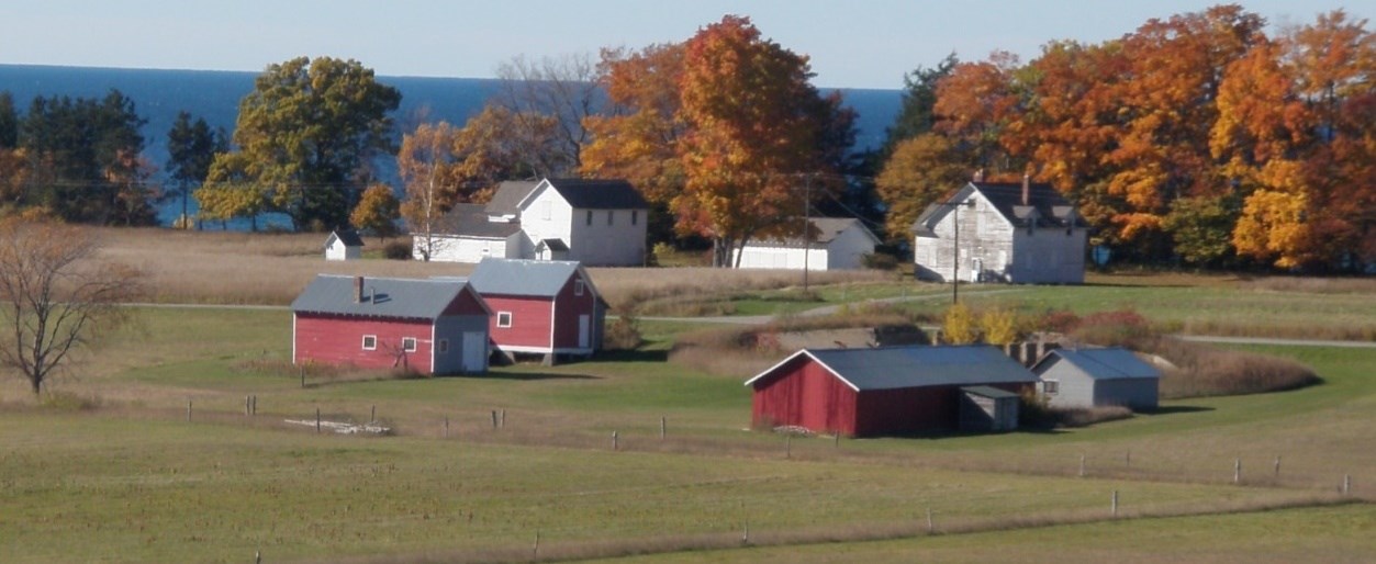 Three white farm buildings sit in front of golden trees and blue water with red outbuildings in the foreground