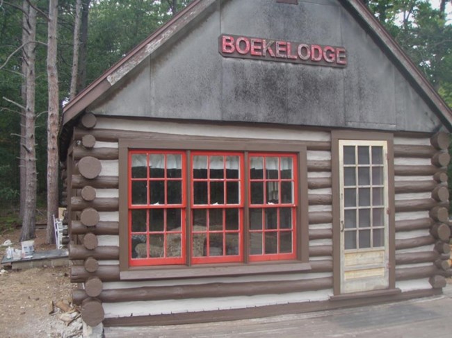 Log cabin with thick, light chinking, red-trimmed windows and red letters that spell out Boekelodge on the peaked roof