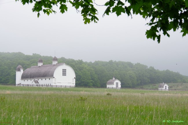 A large, rounded roof barn with two cupolas in the roof and large twelve-paned window and two small outbuildings