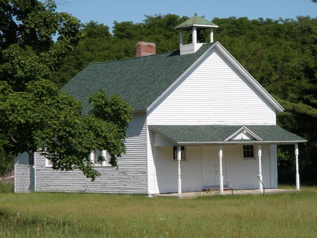 White clapboard building with full front porch and school bell steeple on roof