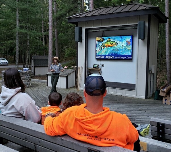 A female park ranger in uniform stands on an outdoor stage with visitors sitting on benches and is talking to the audience in front of a large tv screen with a drawing of a fish on the screen and "Junior Angler with ranger Dana" written.