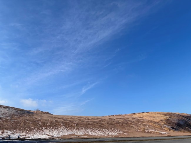 A sand dune in winter under a blue sky with very little snow on the dune.