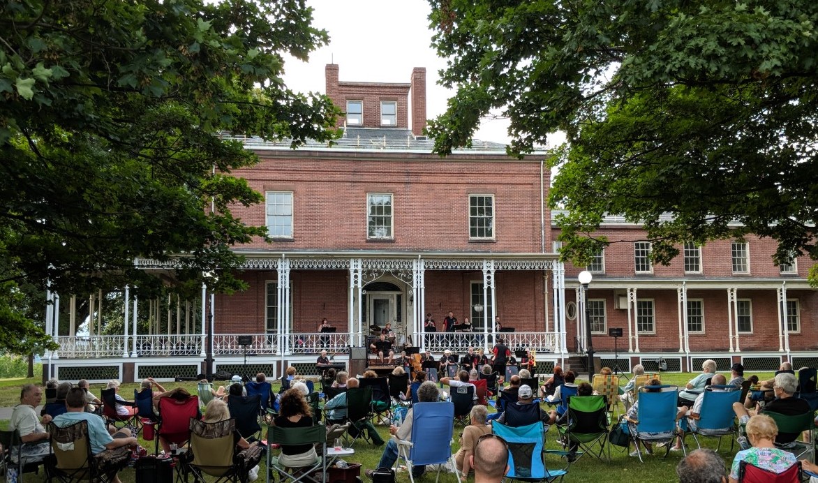 Brick house with white iron railings with 16 band members in black and red polos with saxophones, drums, keyboard and three trumpeters playing.