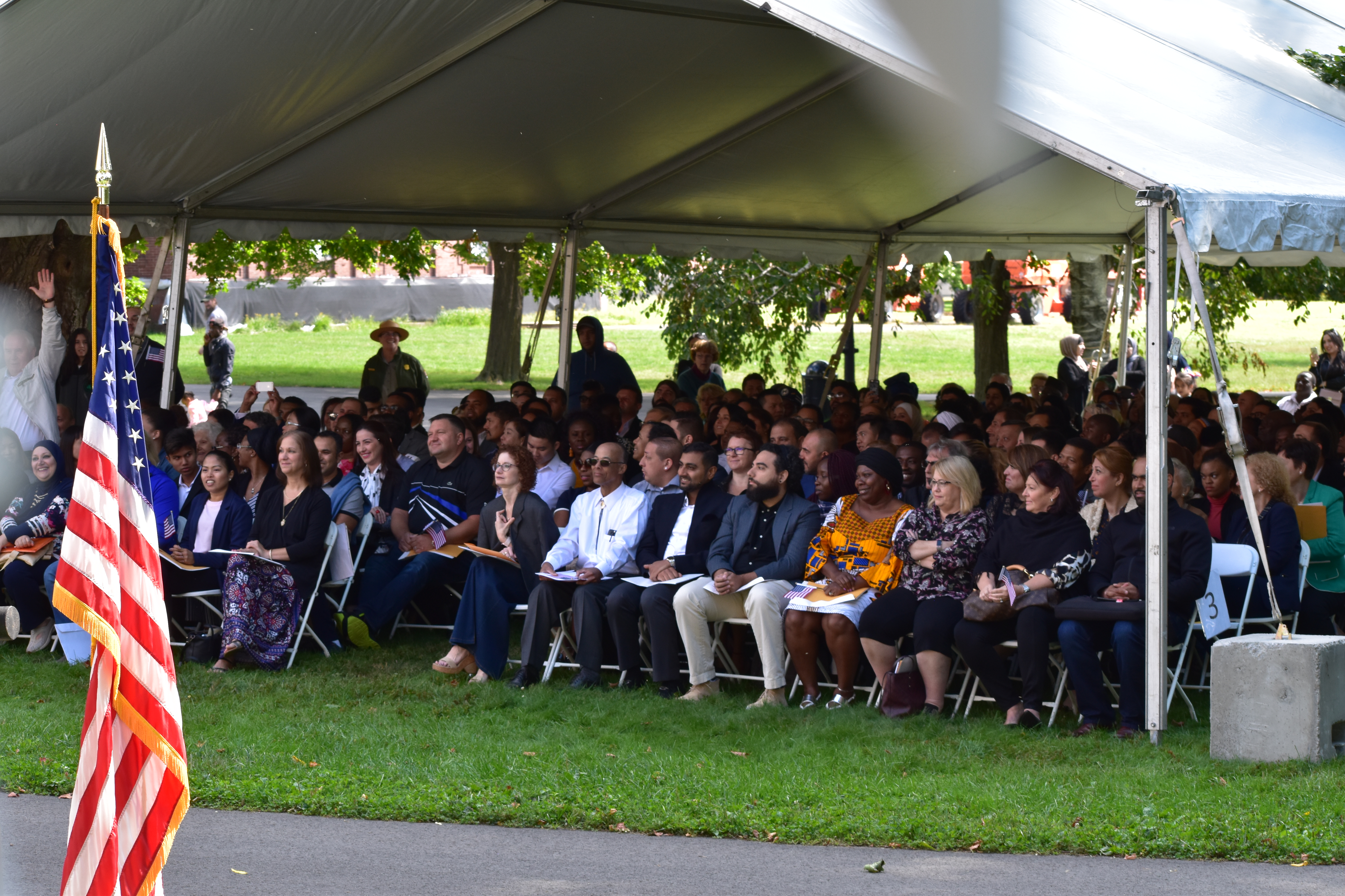 Underneath a white tent, a group of people sit in chairs, with an American Flag in the foreground on the left.