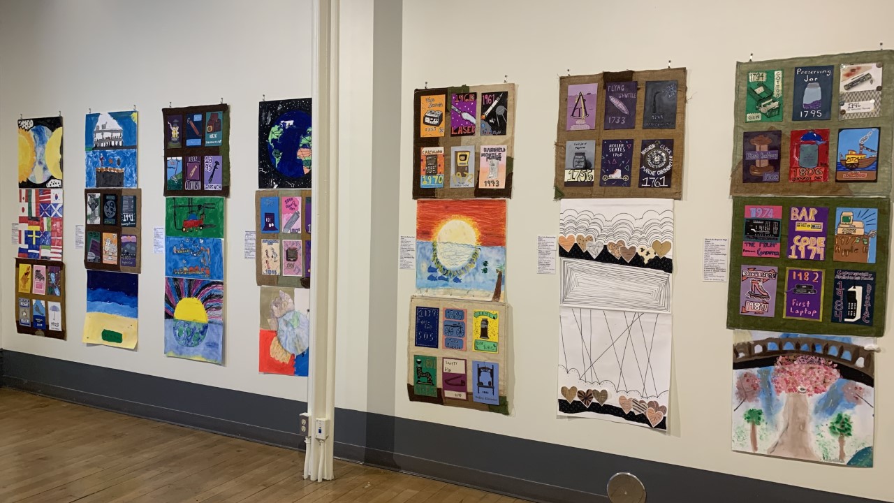 Student artwork on display several 2 x 2 squares of colorful artwork pieces in rows of three hanging on a white wall