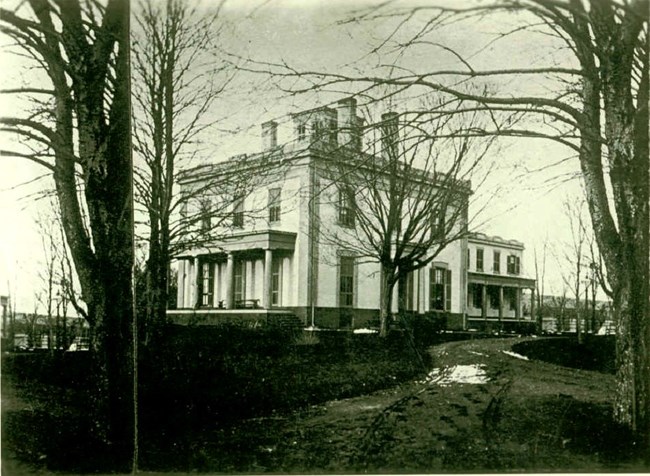 A black and white photo of a multistory building behind bare trees.