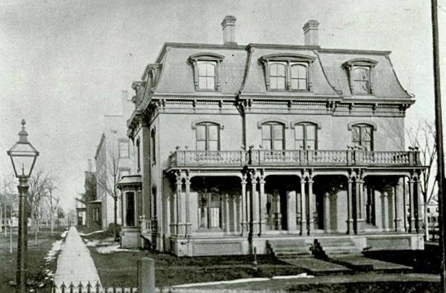 A black and white photo of a multistory building with a porch and balcony. it has multiple windows and a lamp is in the foreground to the left.