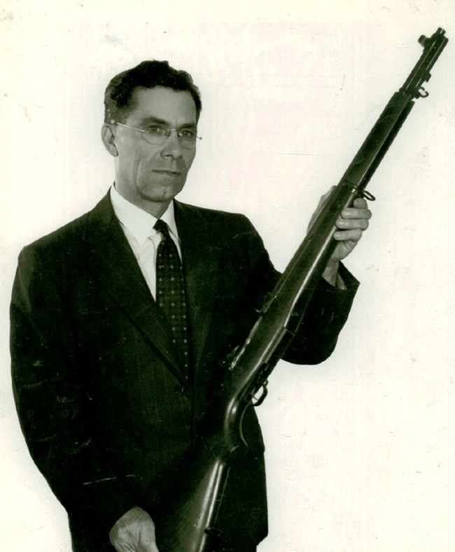 Portrait, half length, of John C. Garand, inventor of the U.S. Rifle, Caliber .30 M1. He wears rimless eyeglasses and dark suit. He holds an M1 at a high angle, left hand just below the forearm swivel and right hand cradling butt and resting on table.