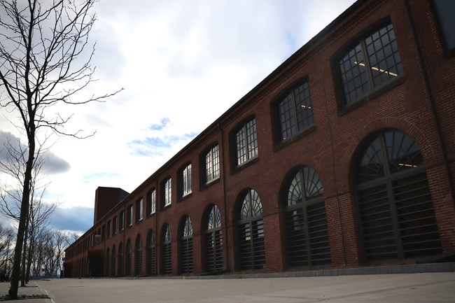 Building 19, a brick building with multiple arched windows reflecting the sunlight. A bare tree is stand to the left of the frame.