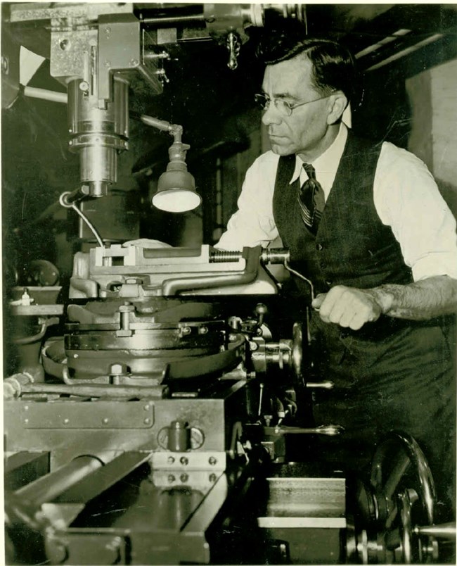 Portrait, three quarter length, of John C. Garand, inventor of the U.S. Rifle, Caliber .30 M1. He wears dark three piece suit sans suitcoat and rimless eyeglasses. He is operating a machine in the Armory Model Shop.