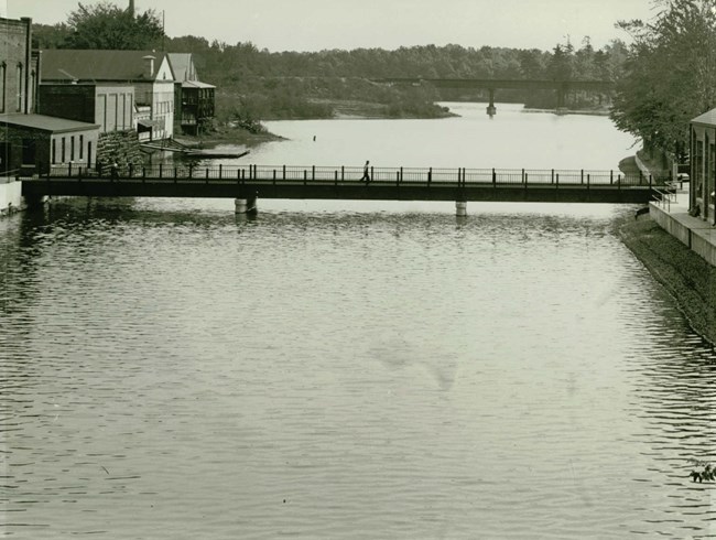 A black and white photo of Watershops Pond with a bridge across pond in middle. Water Shop buildings are on right and left. Beam bridge in background.