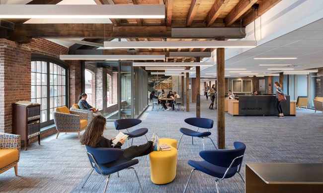A student center, with students walking around and sitting at tables studying. The building blends rustic and new in it's interior design.