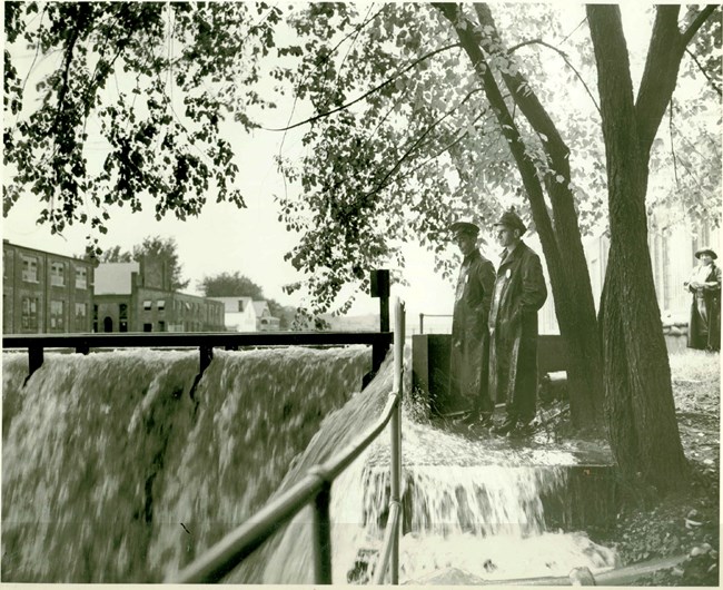 A black and white photo of two men standing in rain coats next to a damn that is flooding with water rushing over the top.