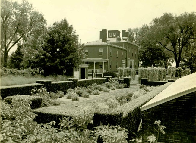 A black and white photo of a building set behind gardens and hedges.