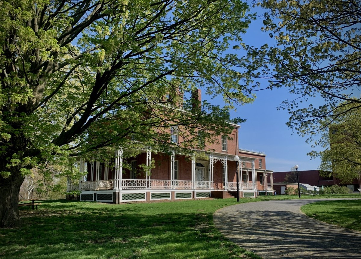 A multistory red brick building with a covered porch. The building sits behind trees on a spring day.