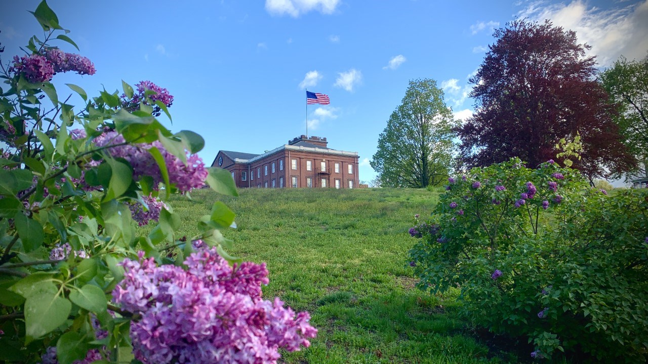 Up on a hill sits the Main Arsenal in early spring with the flag flying from atop the clock tower. Lilacs are in bloom in the foreground.