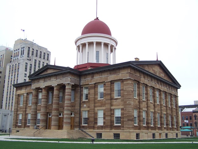 A multi-story, tan stone capitol building with many windows and large columns in front. The top has a large red dome.