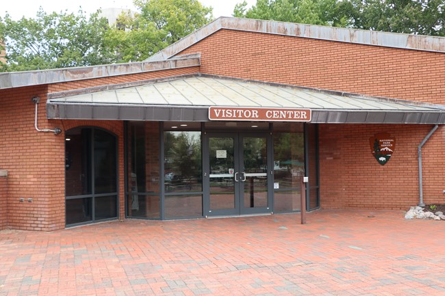A one-story, brick building with the National Park Service arrowhead on one wall and a "Visitor Center" sign above glass doors.