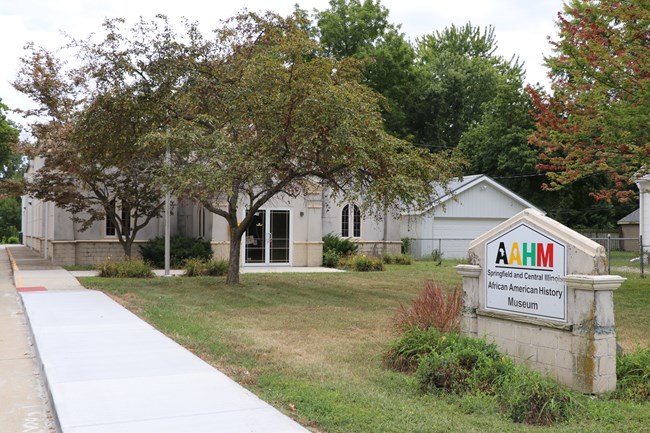 A small, light colored building hidden behind some trees with a sign in front for the "Springfield and Central Illinois African American History Museum".