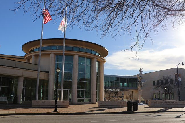 A large, circular building with a glass front, stone columns, and two flag poles in front.