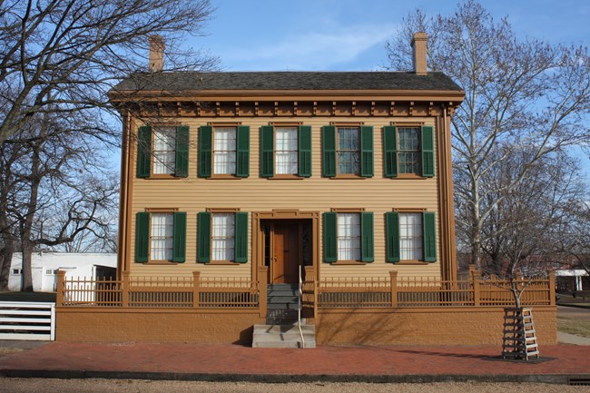 A two-story, tan home with green shutters and a brown fence in front with a brick walkway.