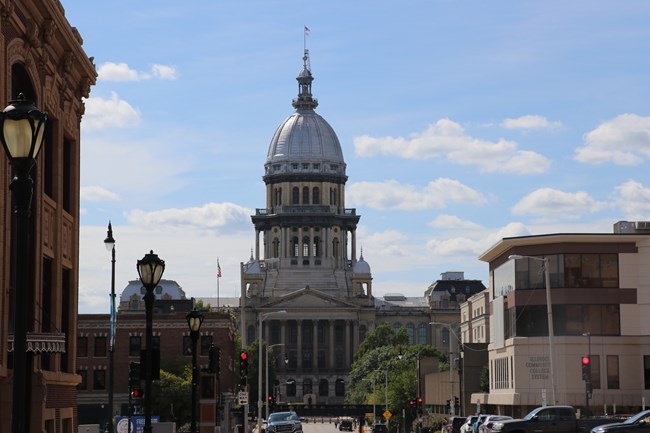 A large, domed capitol building with a flag on top, sits at the end of a street lined with buildings.
