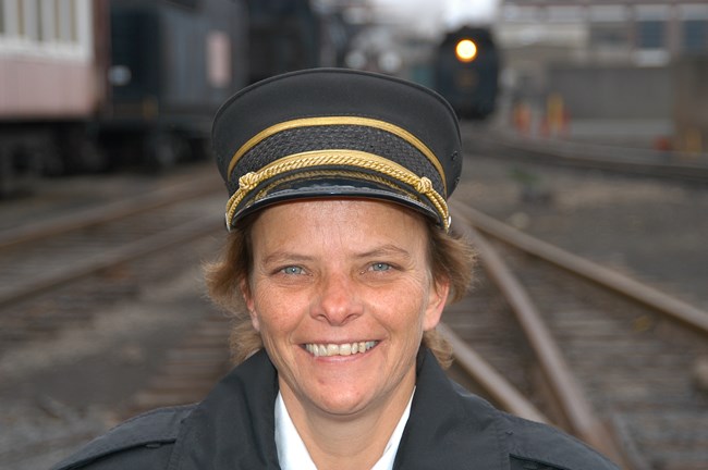 A portrait photograph of Lori Staely in a black and gold conductor's uniform. A railyard is blurred in the background with a steam locomotive's rear light lit near the upper right corner of the photo.