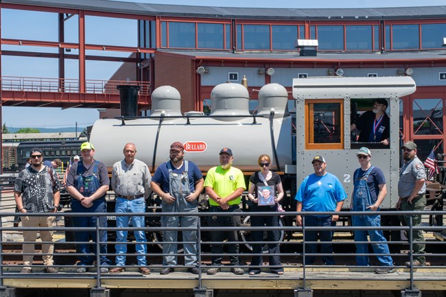 A group of volunteers pose with a small black and gray locomotive on turntable