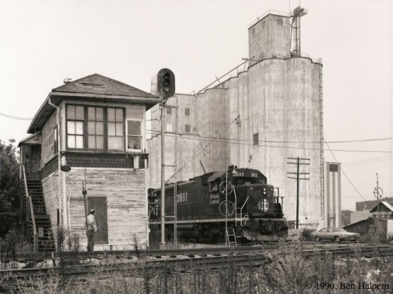 A black and white photo of a diesel-electric locomotive passing a switch tower with grain elevators in the background.