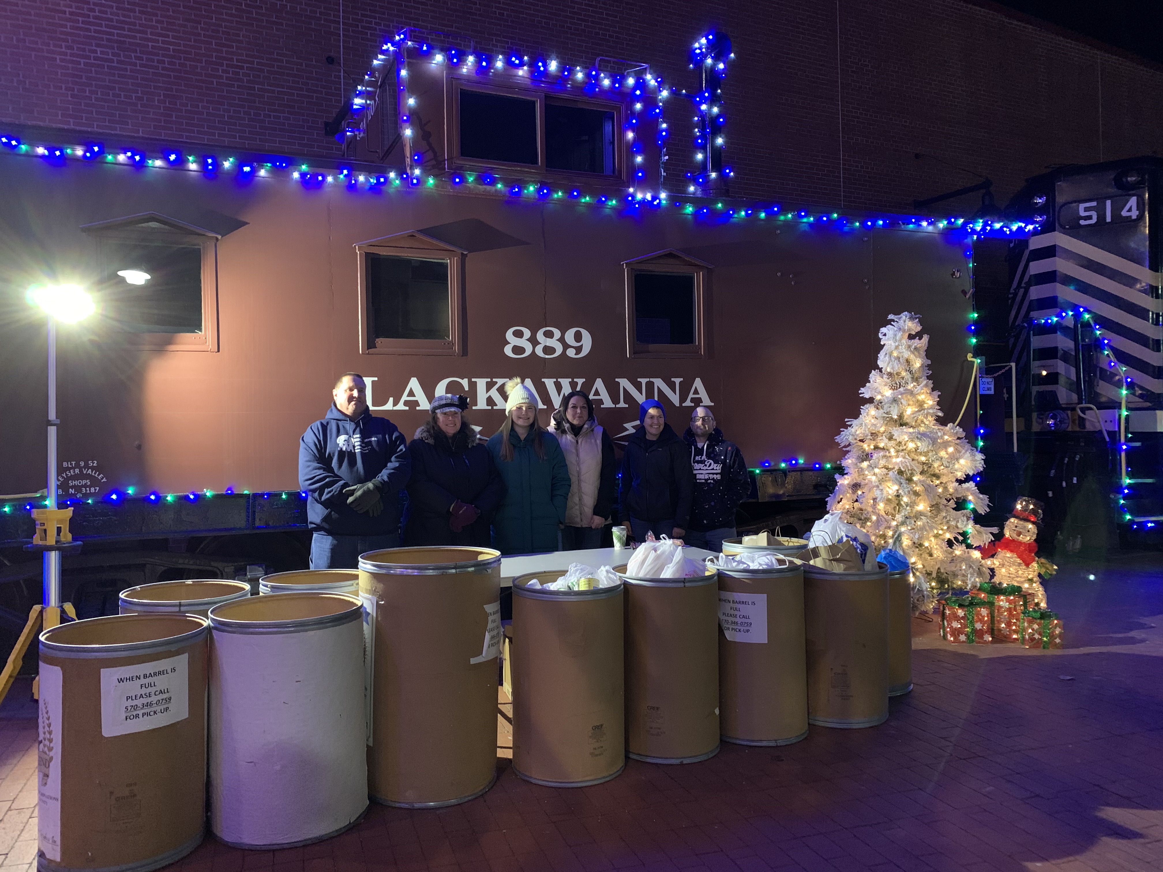 A group of volunteers pose behind a table of food donations, the atmosphere around them is full of winter holiday decorations