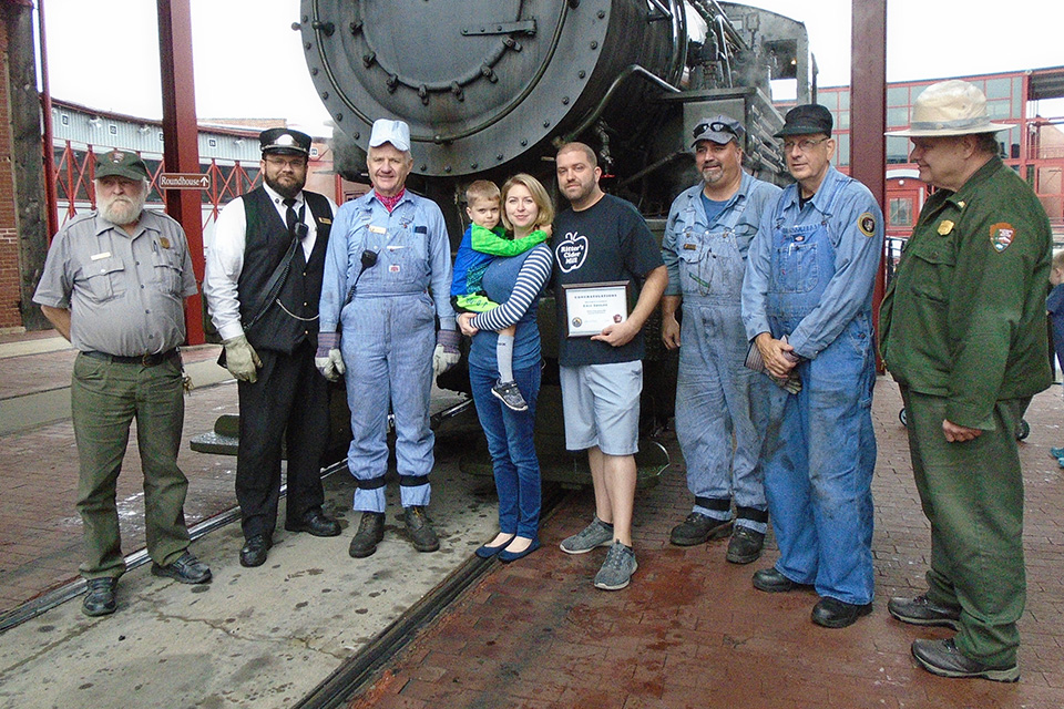 Railfest 2018 Chugga - Shulde Family and Crew 9 adults stand in front of a black iron steam engine. A young boy is in the arms of a woman in the middle of the row.