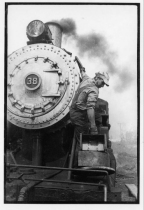 A black and white image of a man in work clothes maintains a steam locomotive near it's smokebox.