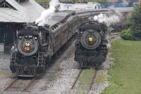 two steam locomotives, both pulling passenger trains, pass the loading platform at Steamtown