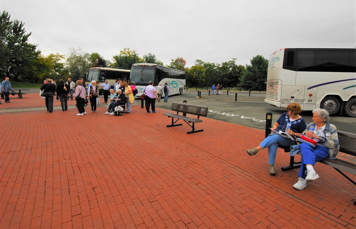 Three tour buses in a parking lot with elderly visitors waiting nearby