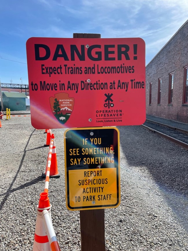 Two signs on a post. Red sign on top announces danger and to expect trains at any time. Yellow sign below encourages visitors if they see something, to say something.