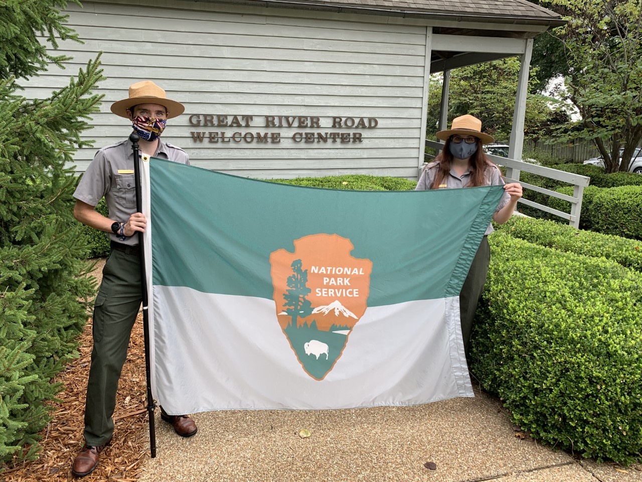 Two park rangers hold a grey and green flag with an arrowhead national park symbol in front of a building.