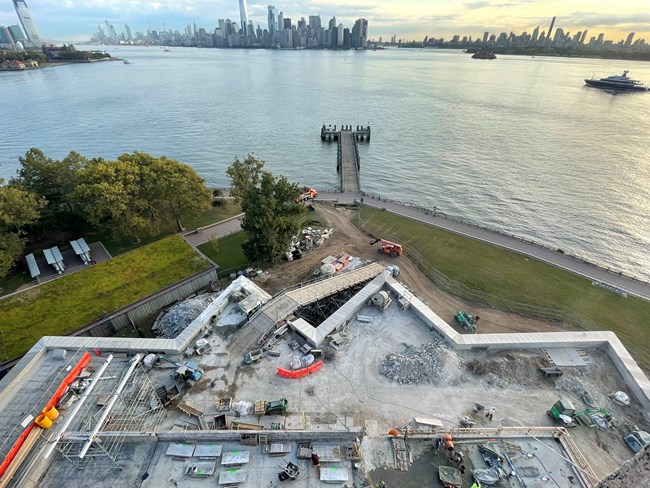 Construction is seen on happening on the top of the Fort Wood level. The area is baricaded off and has construction equipment. The NYC harbor is seen in the background.