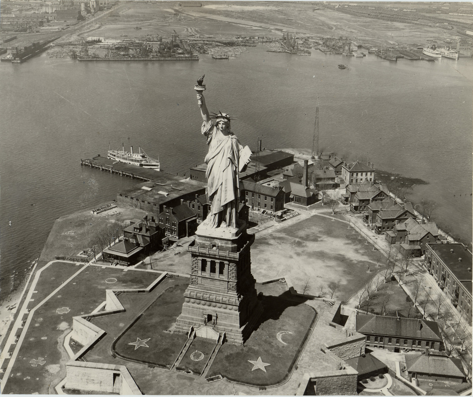 An aerial photograph of an island. The Statue of Liberty is standing on the front of the island with various buildings on the backside.