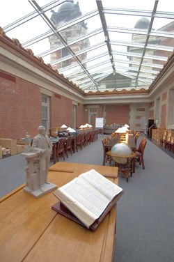 An open book on a table in the foreground, dark wood desks in the rear, brick walls and clear glass roof above.  The Reading Room in the Bob Hope Memorial Library.
