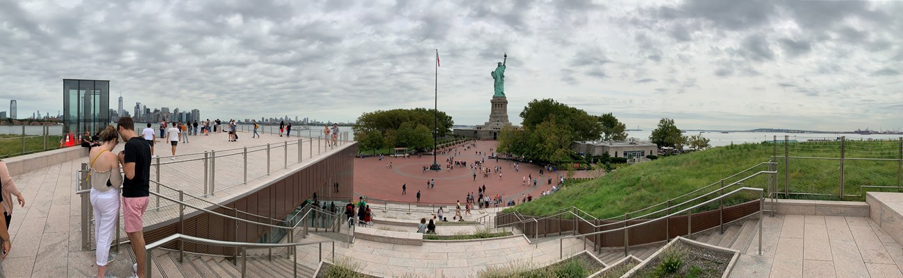 A photo taken from the top of the museum shows the New York City skyline to the left of the image, the Statue of Liberty centered in the distance and visitors around the island.