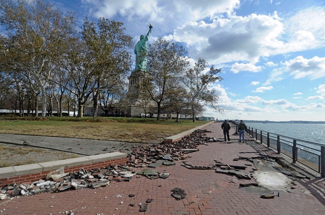 Two people walk around Liberty Island with bricks displaced and tree limbs down.