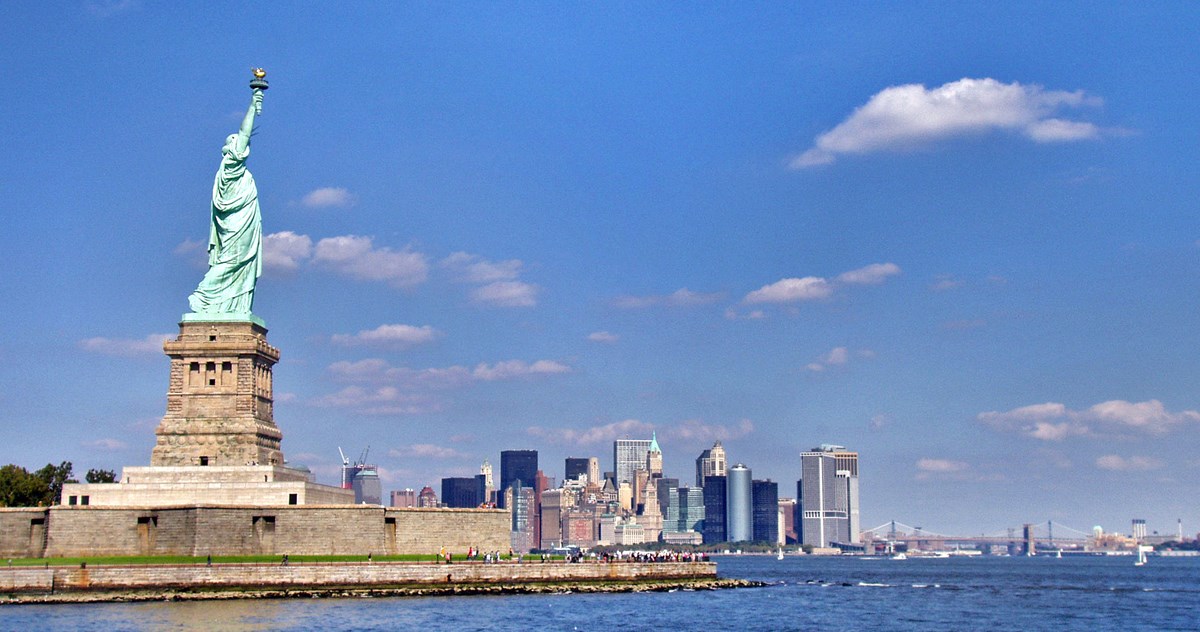 The Statue of Liberty stands against the Manhattan skyline and a blue sky.