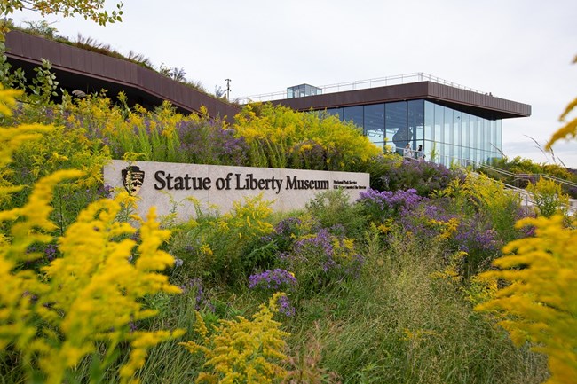 The exterior of the museum with native purple and yellow plants surrounding the sign which reads: "Statue of Liberty Museum, National Park Service, U.S. Dept of the Interior" with an NPS arrowhead.