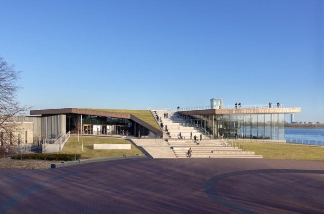 Statue of Liberty Museum as seen from the grounds of Liberty Island. The building has floor to ceiling windows, a green roof and steps in the center of the building to the roof.