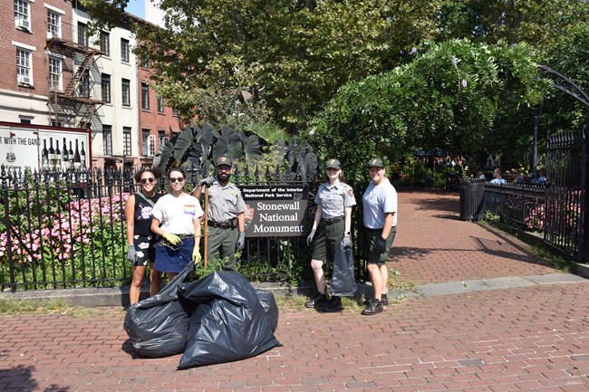 5 people standing in front of the Stonewall National Monument sign at Christopher Park. The two people on the left are wearing casual t-shirts while the other three are in green pants and grey shirts with national park service hats.