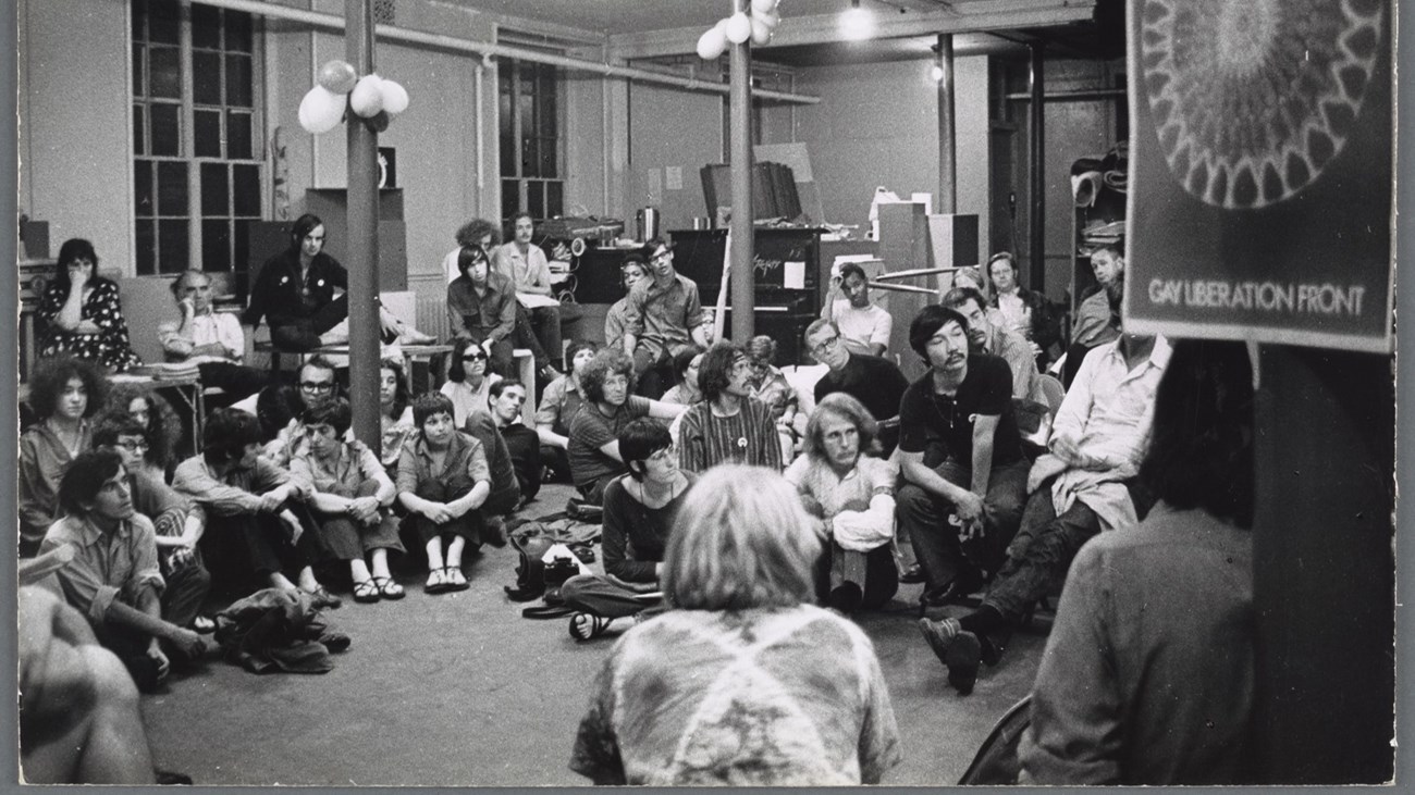 Many people sitting in chairs and on the floor with a Gay Liberation Front (GLF) sign in the foreground