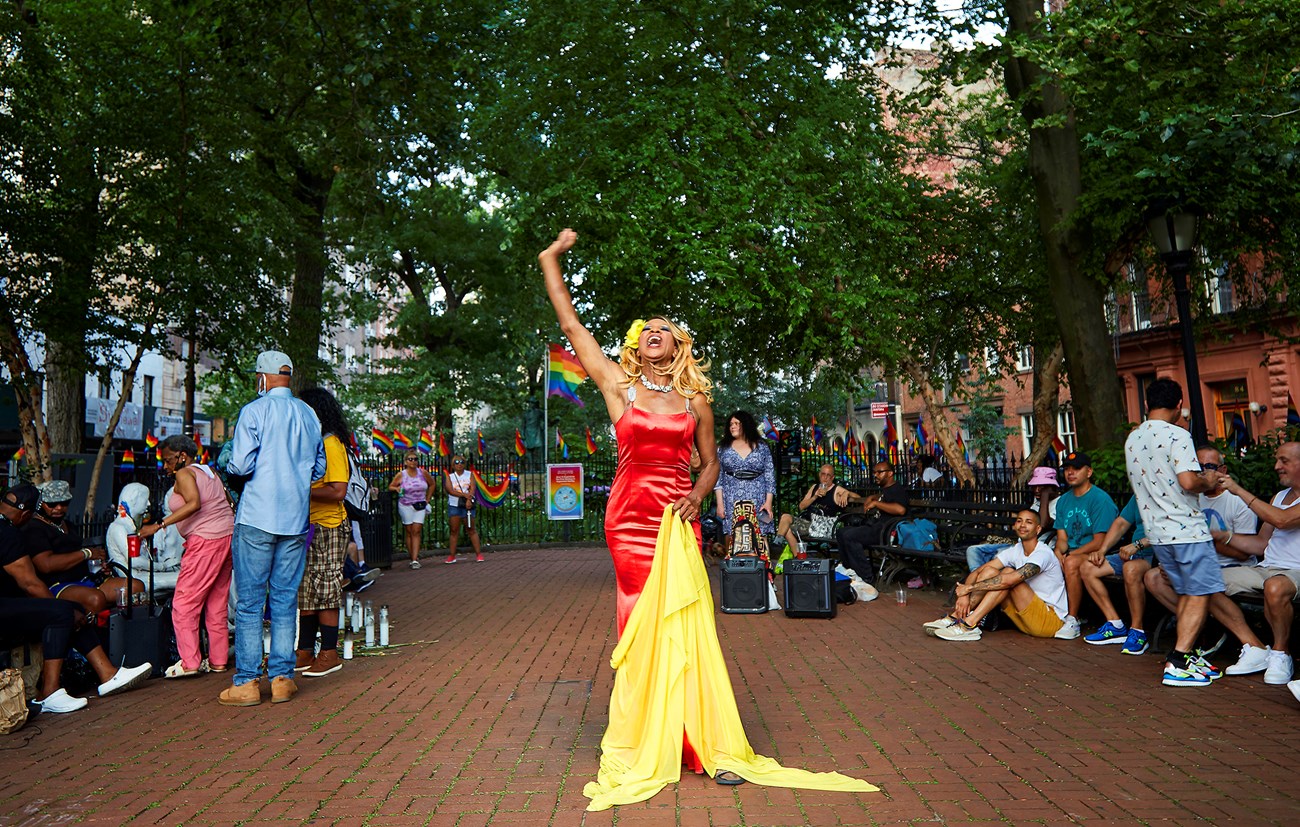 Portrait of Miss Simone at the Stonewall Monument, Miss Simone standing in Christopher Park holding yellow fabric with many people looking at her